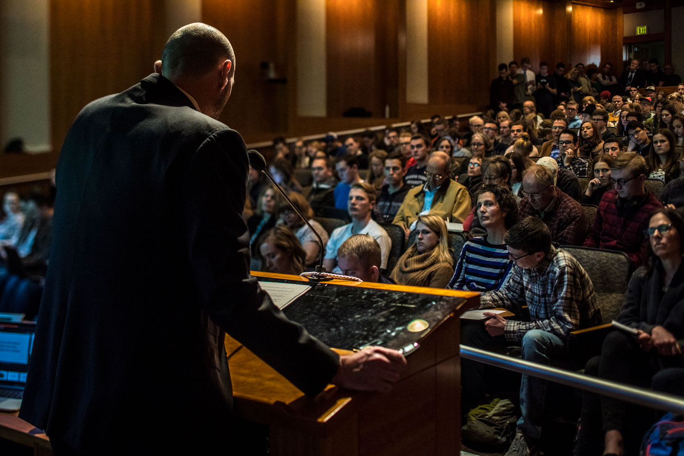 Back view of Adam Miller, standing at a podium, facing a large crowd of students.