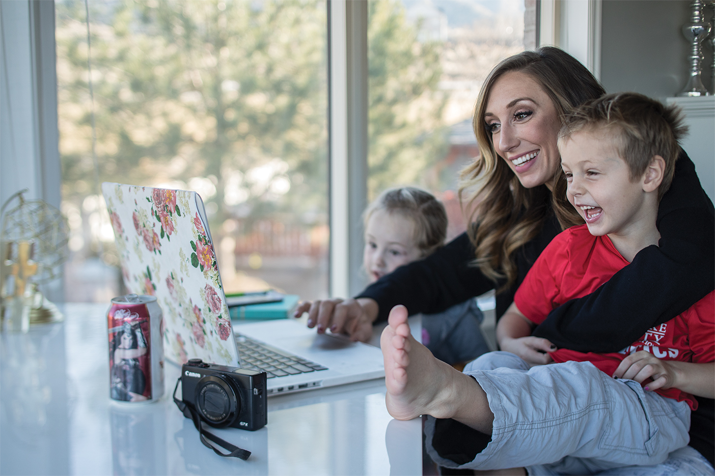 Jordan page sits at her table with one of her kids next to her and one on her lap as she looks at something on her laptop screen.