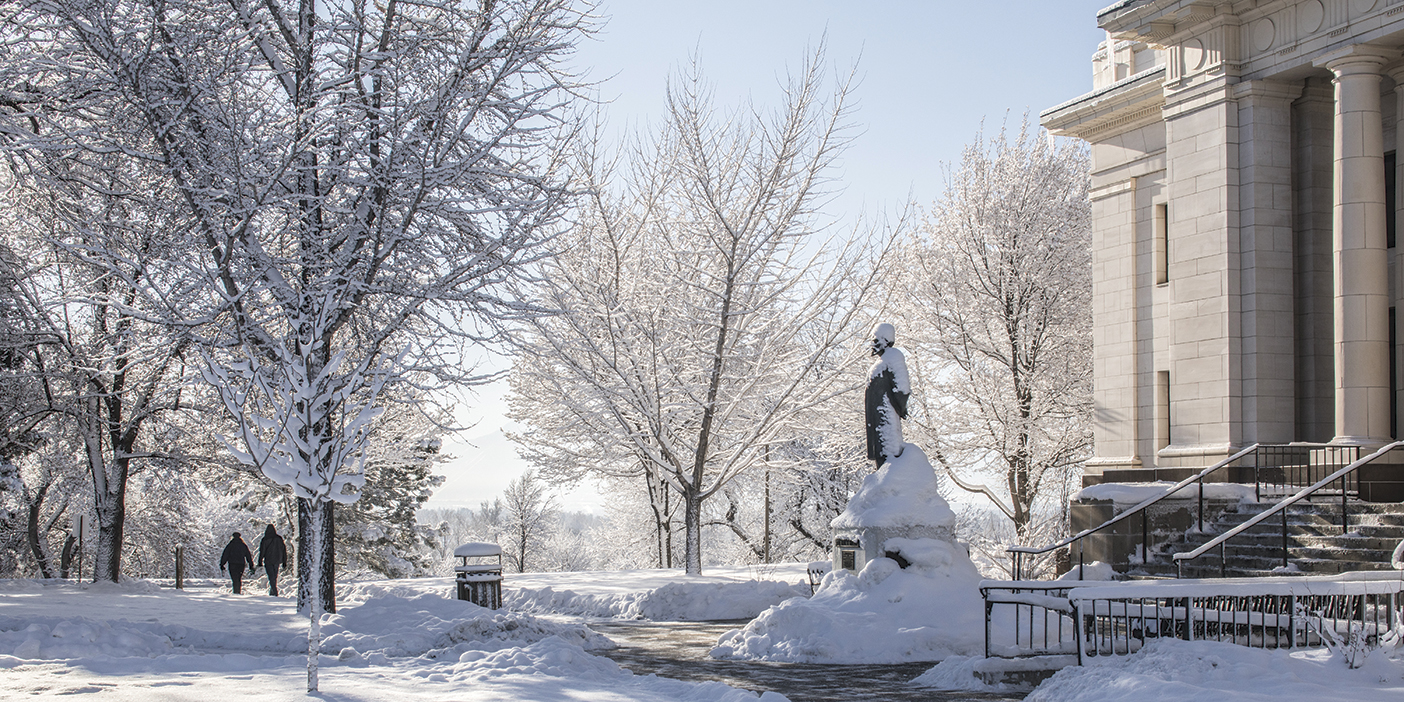 A photo of snow covered trees and the Karl G. Maeser statue covered in snow outside the Maeser building on BYU campus.