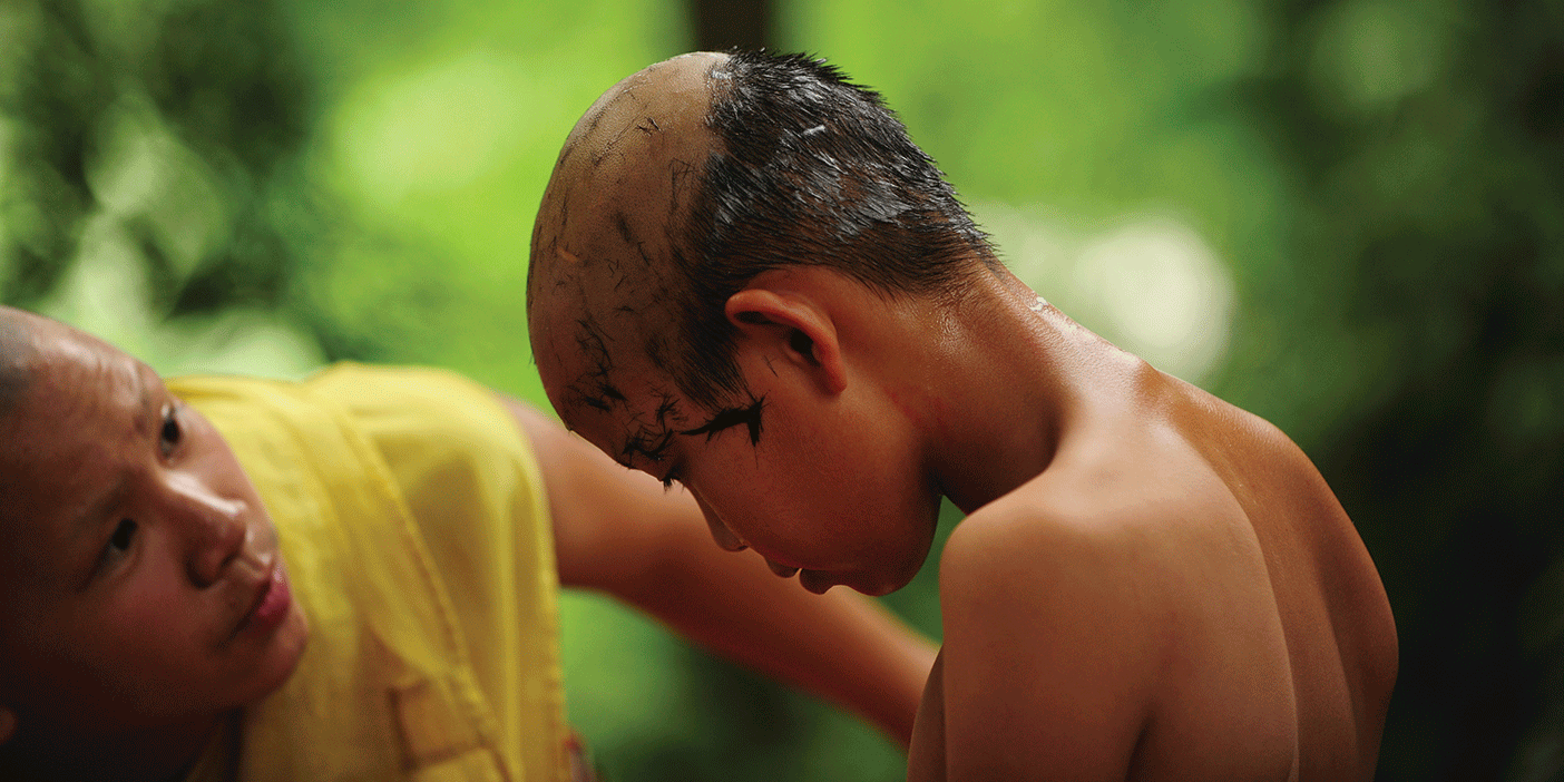 A young Buddhist monk bowing his head down while another monk shaves his head.
