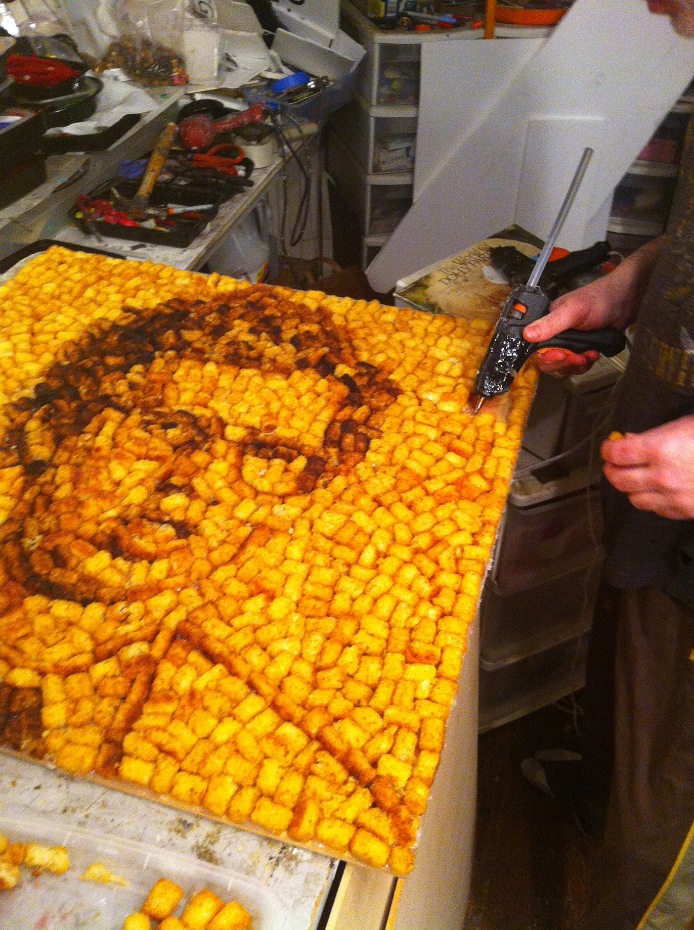 Photo from the process of making the cover art showing Napoleon Dynamite made out of tater tots. This photo shows the Mecier gluing on the finishing touches.