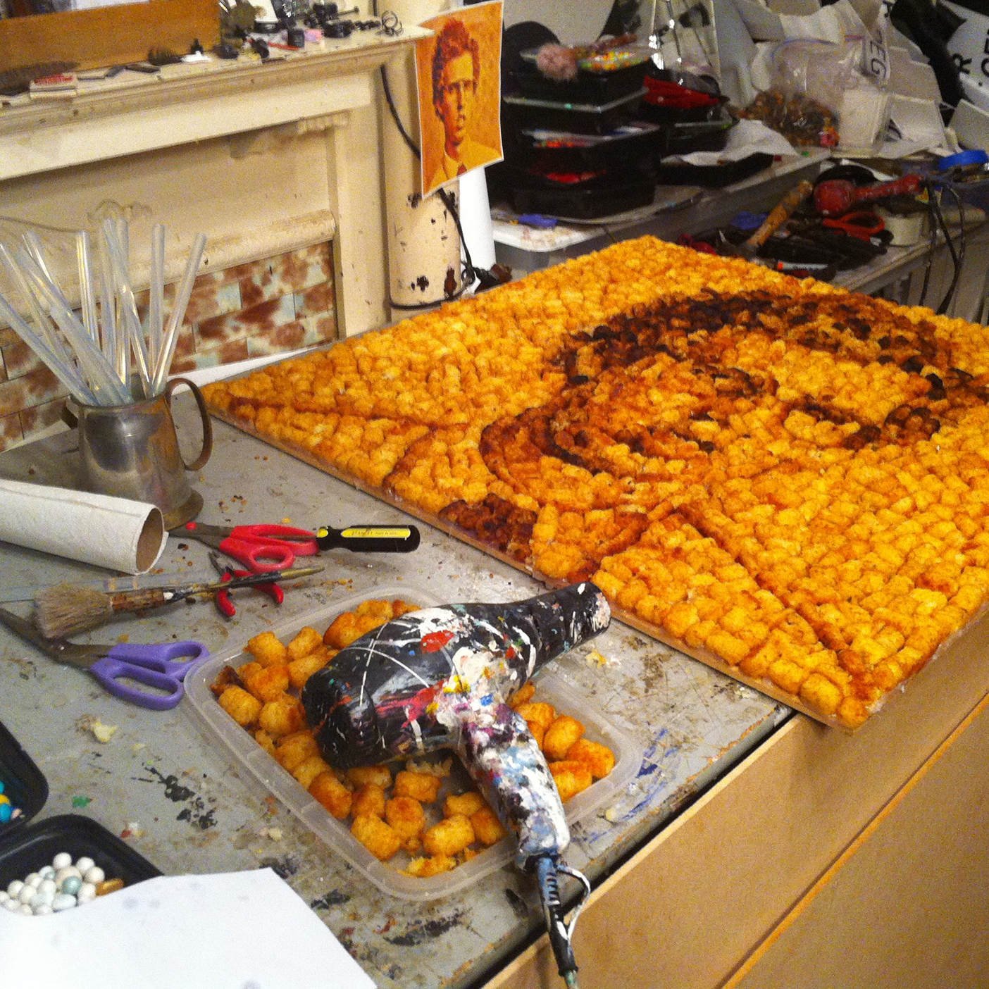 Photo from the process of making the cover art showing Napoleon Dynamite made out of tater tots. This shot shows the final image in the background and hot-glue guns and loose tots in the foreground.