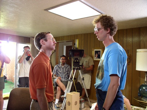 Kip and Napoleon are in the foreground, filming a scene in their home. In the background, you can see the video camera and the production crew.