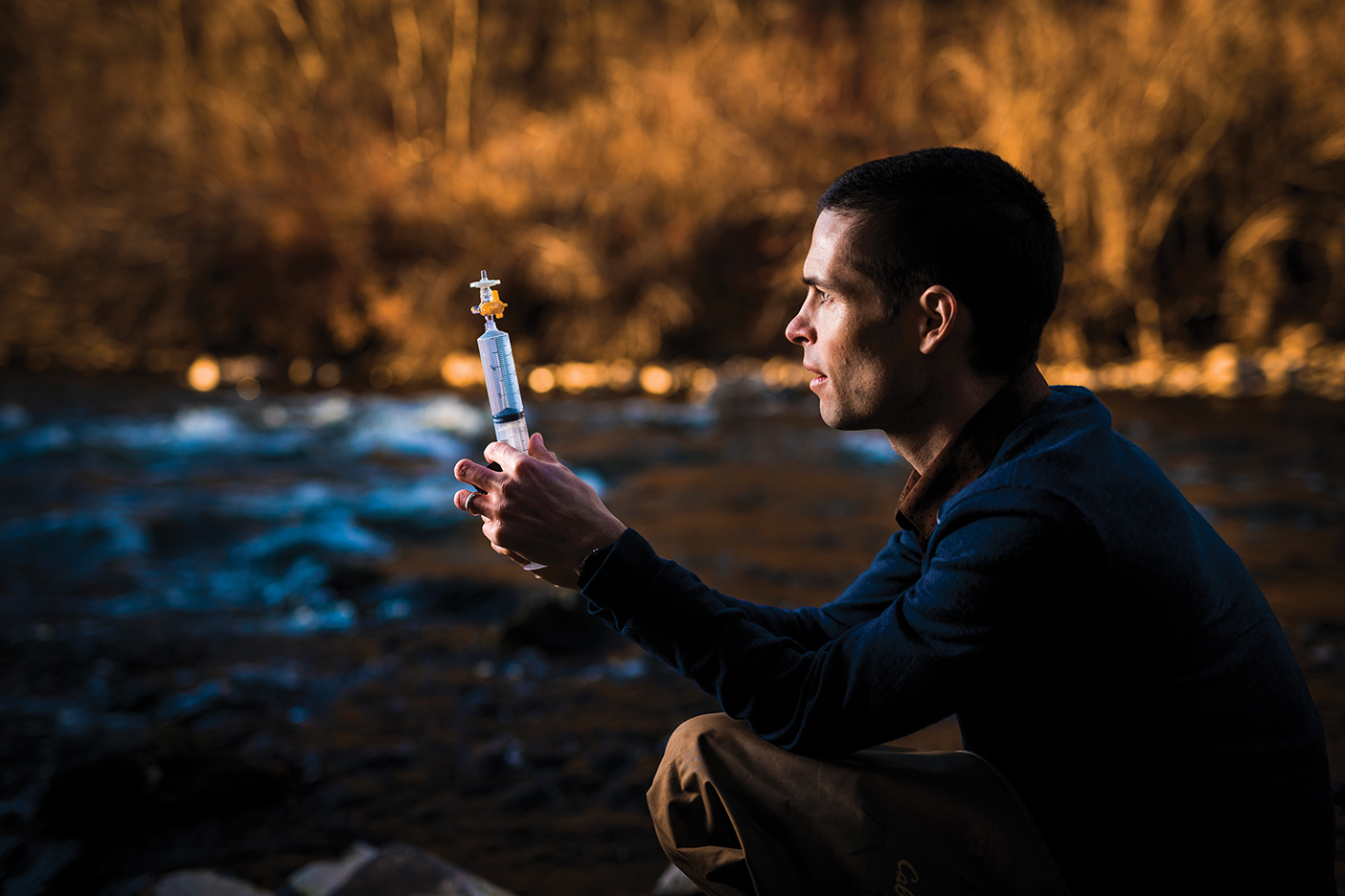 BYU professor Ben Abbott looking at a syringe, crouched in a stream.