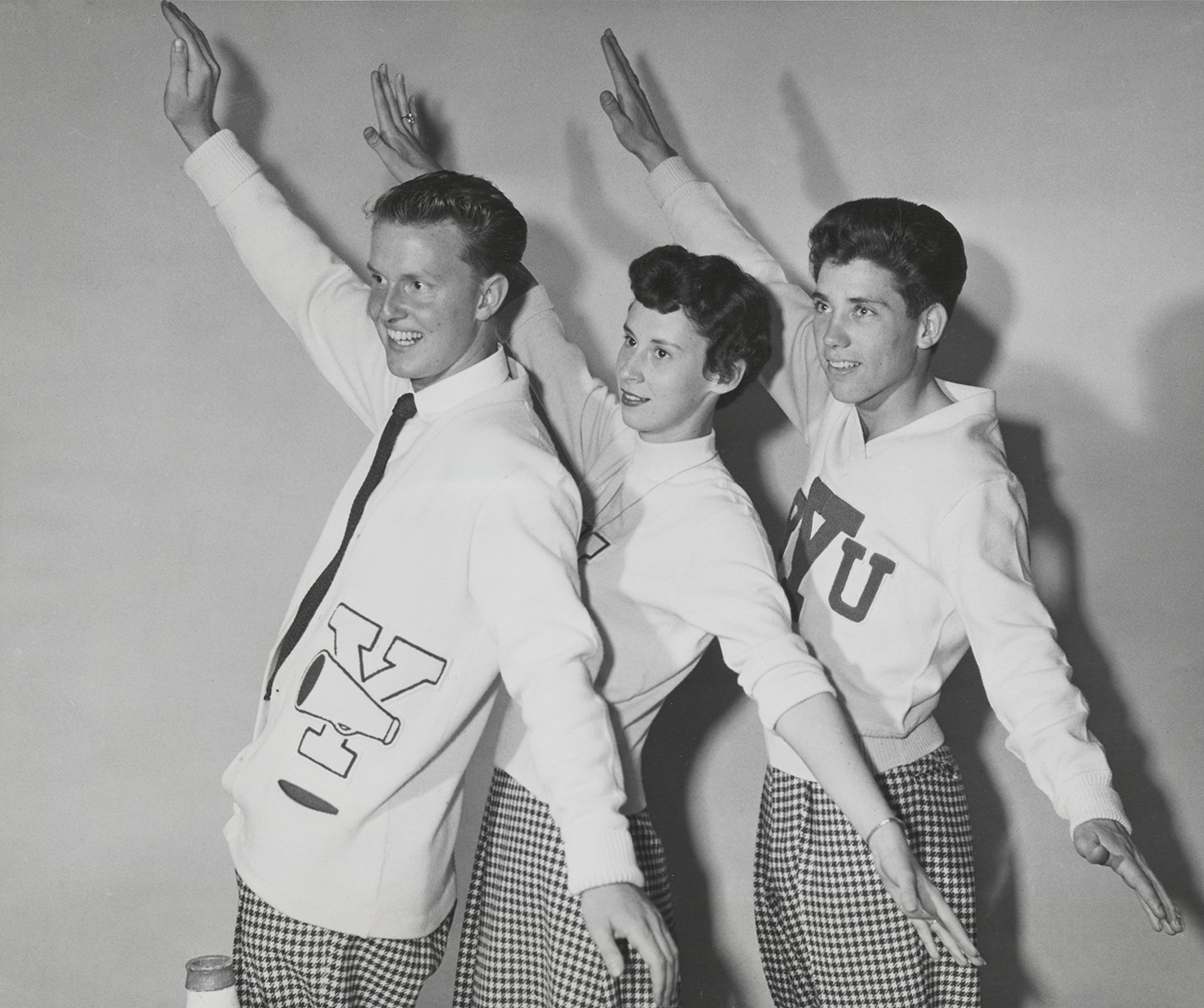 A black and white photo of three cheerleaders from 1954 raising their arms.