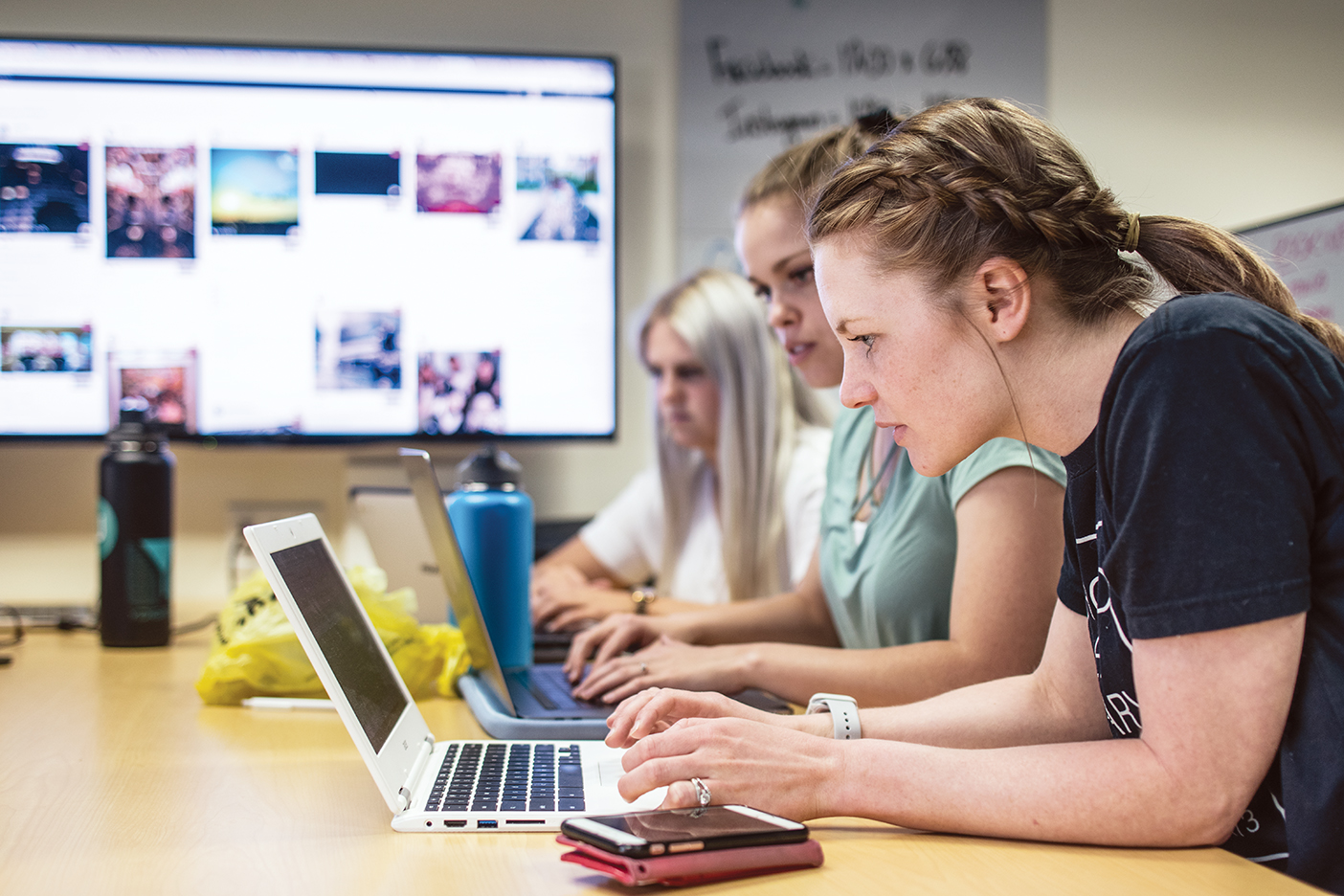 Several Y Digital students type on their laptop computers while a large screen mounted on the wall behind them displays a web page.