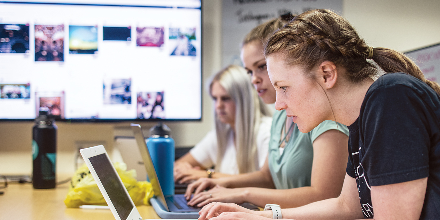 Several Y Digital students type on their laptop computers while a large screen mounted on the wall behind them displays a web page.