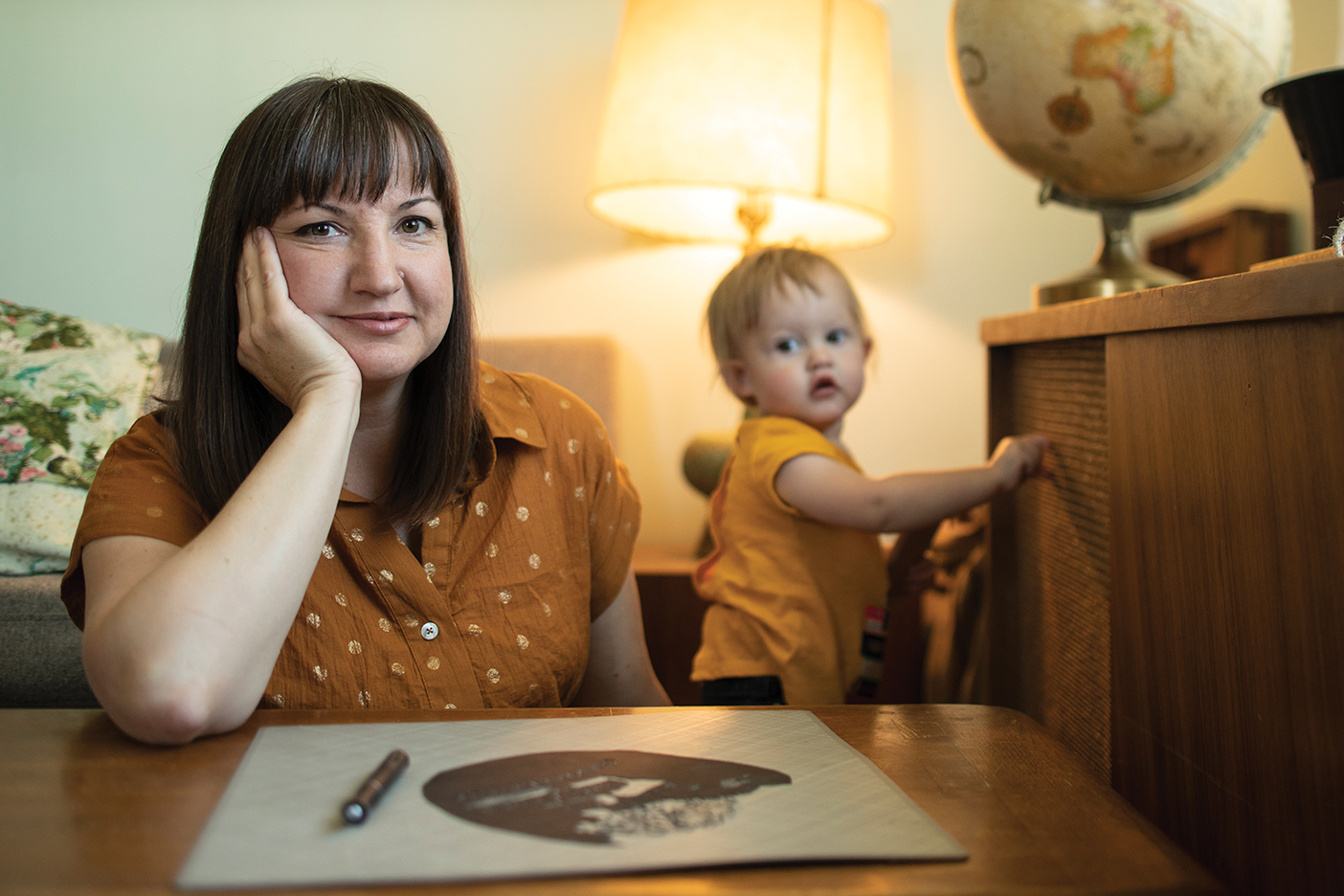 Her daughter playing in the background, Cindy Bean sits at a desk with one of her paper-cut creations.