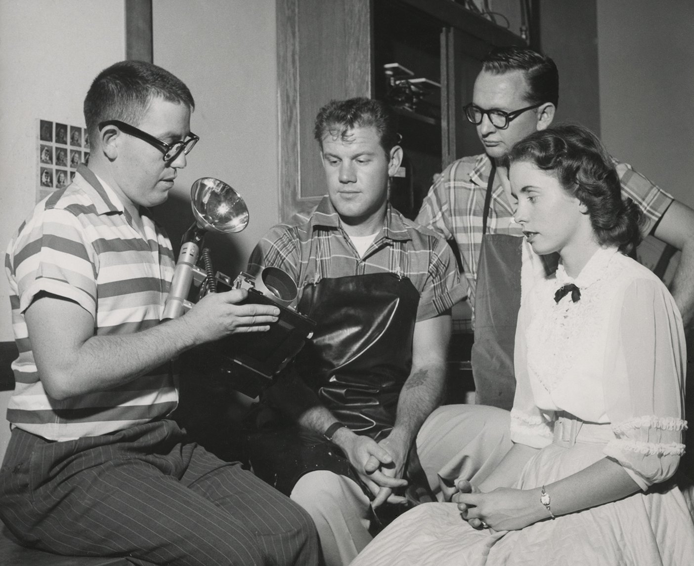 Three students gather around an instructor, who teaches the students how to use a camera and flash in 1957.