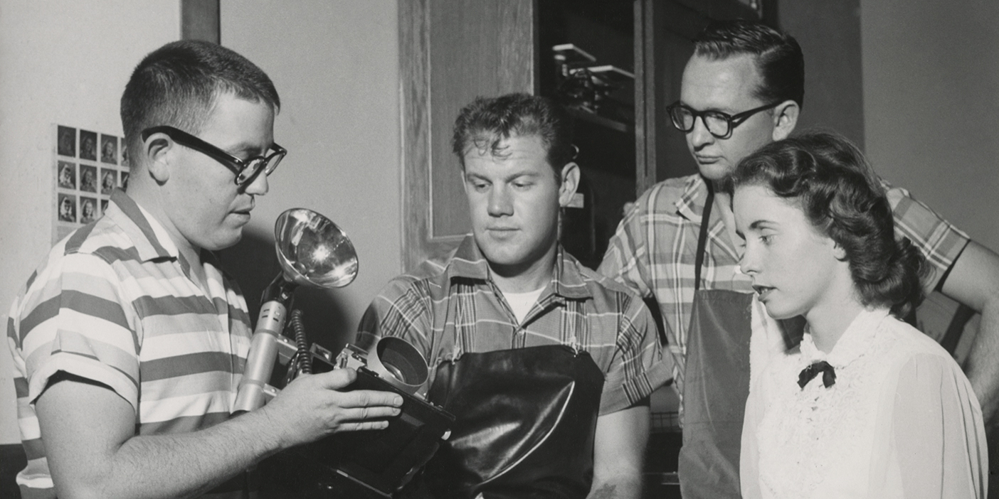 Three BYU students watch as their instructor demonstrates how to use a camera and flash in a 1957 photojournalism class.
