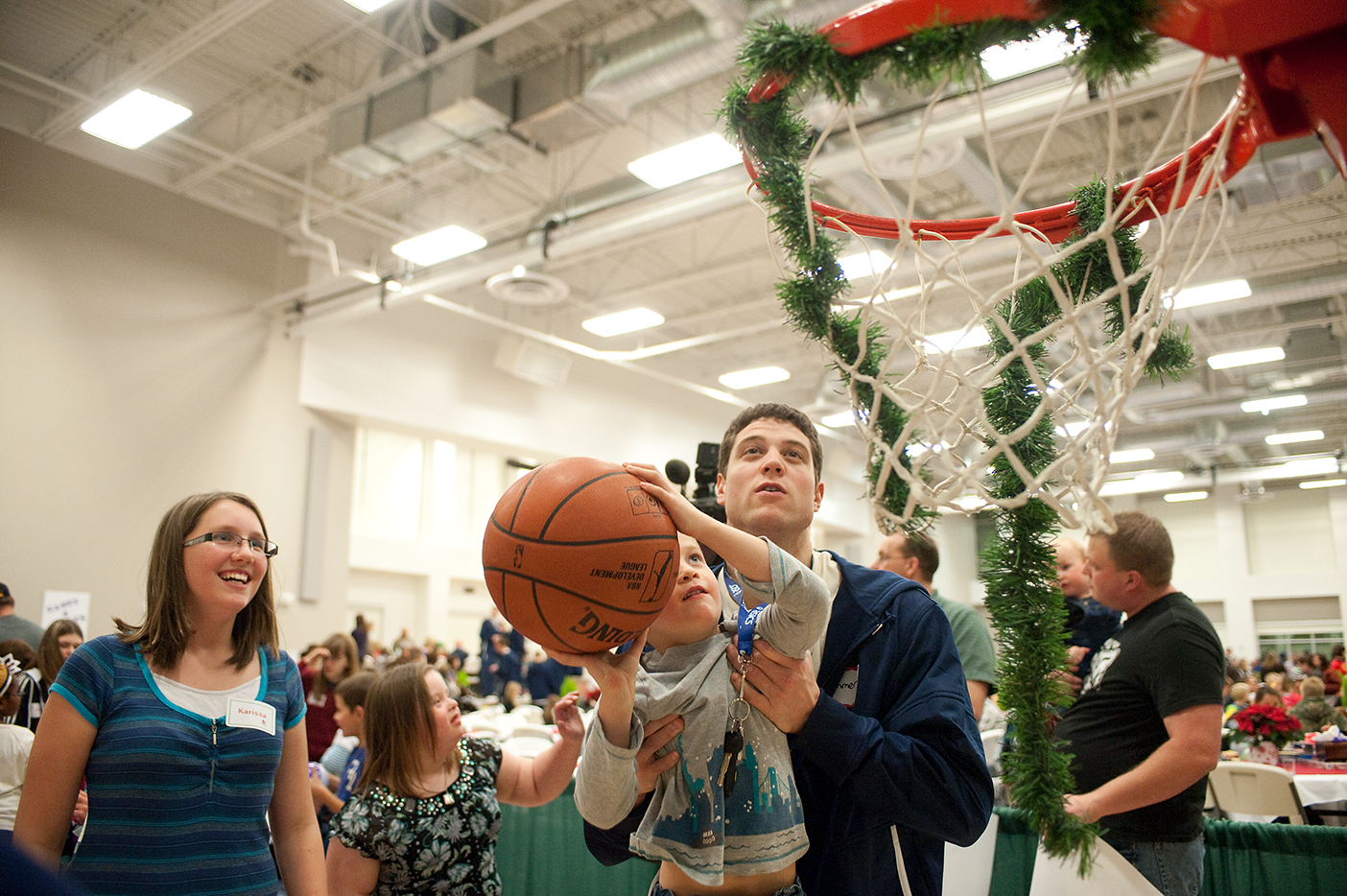 Jimmer Fredette lifts up a small boy to make a shot in a garland-decorated hoop as a woman watches.