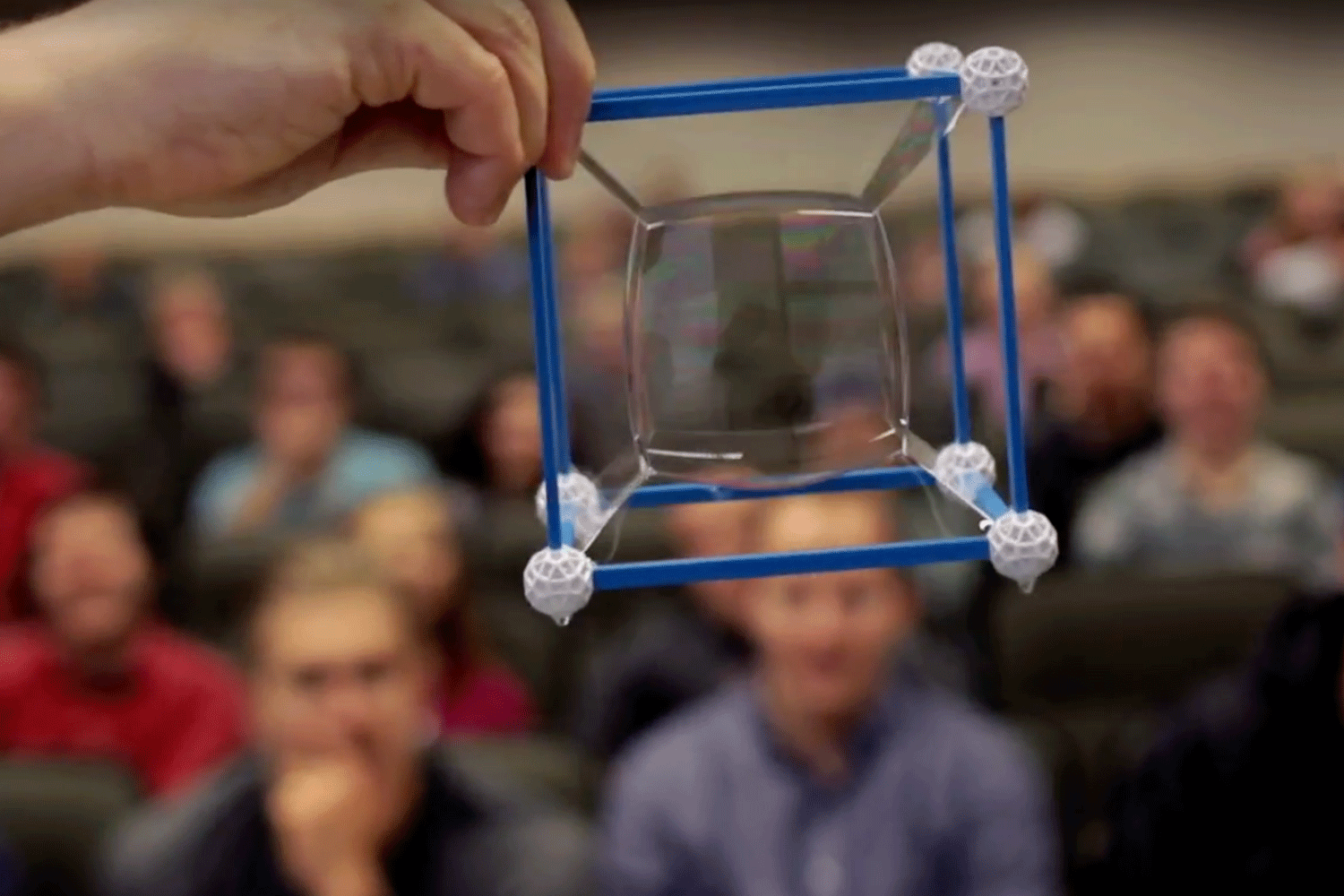 A screenshot from our Micahel Dorff "Thing of Beauty" video on YouTube. The screenshot shows Dorff's outstretched arm holding a cube made of straws. He has dipped the straw cube in a solution of bubbles twice, creating a cube within a cube.