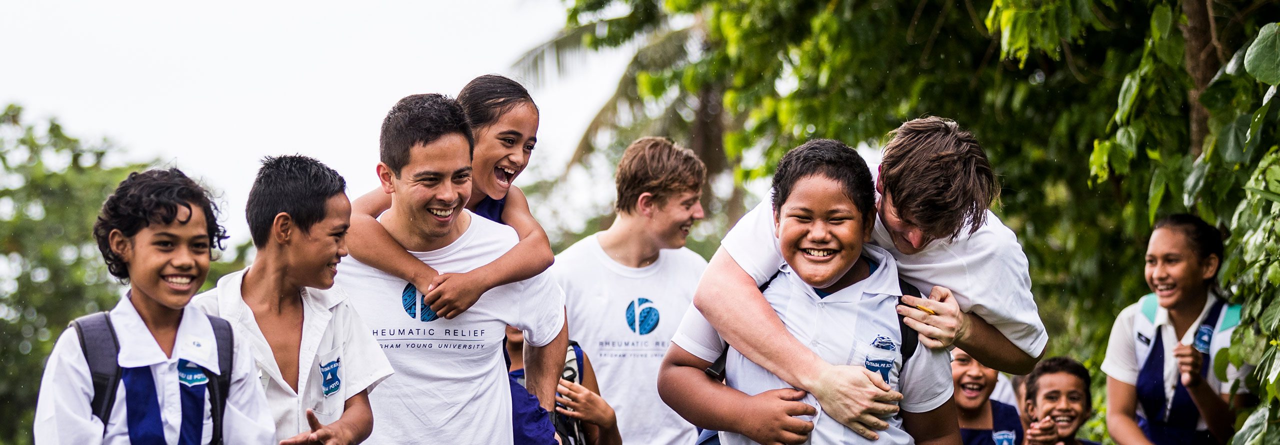 Young Samoan students interact and have a good time with a group of the BYU's relief team as they walk. Palm trees are in the background.