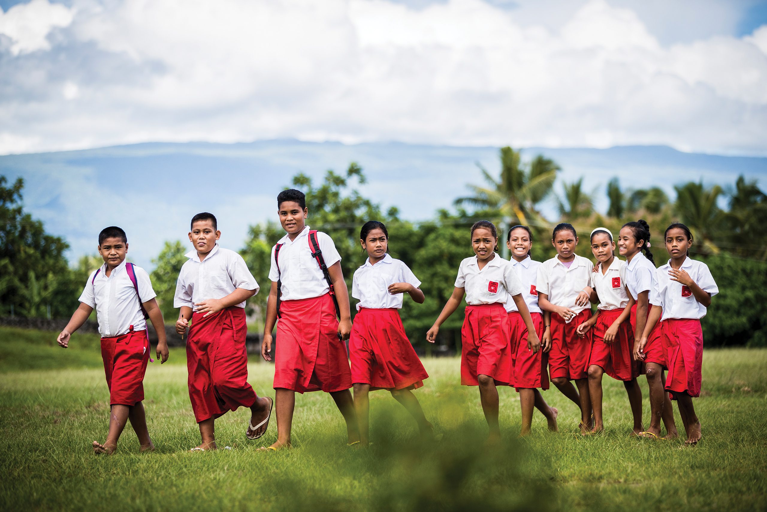 A group of young Samoan students walking across a field with palm trees in the background.