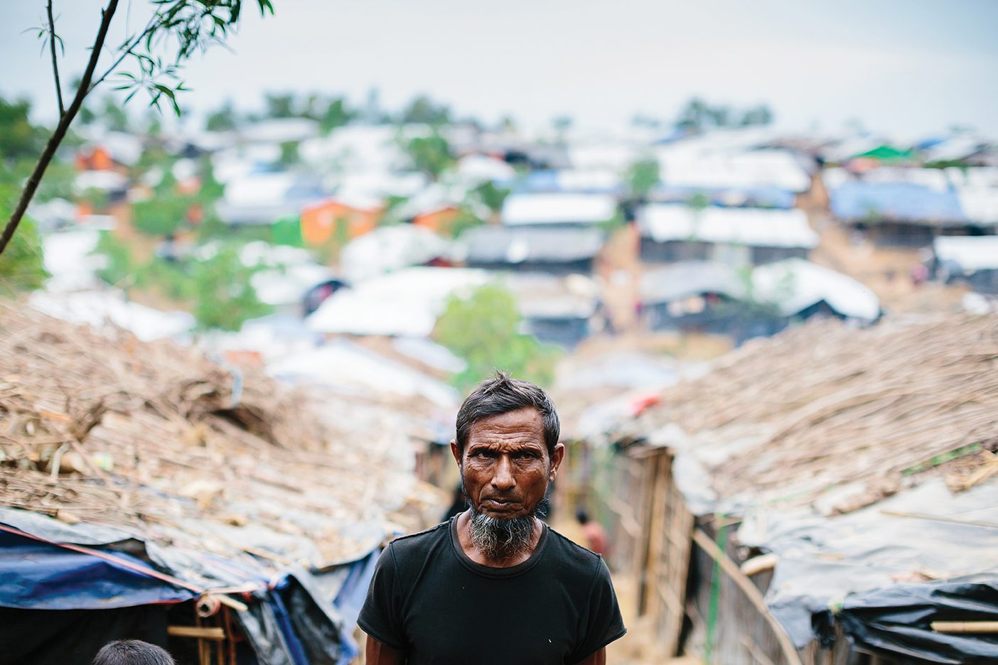 A Rohingya man walks in between grass huts in a refugee camp.
