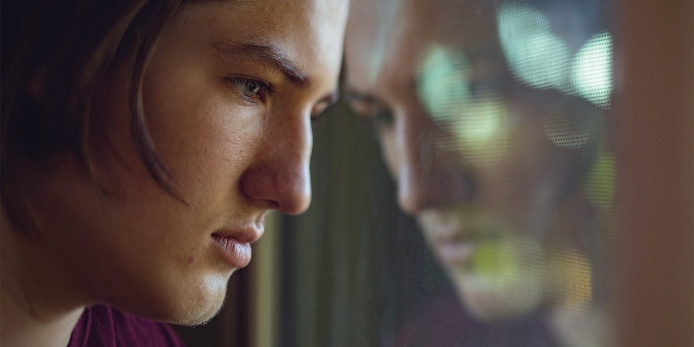 A despondent teen leans against his reflection in a window pane.