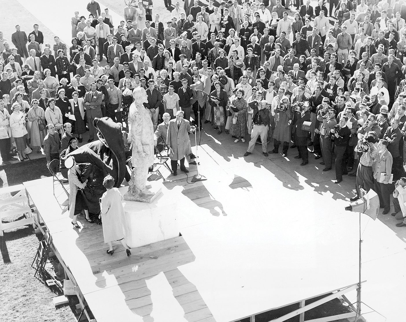 An old photo of the unveiling of the Karl G. Maeser statue placed outside of a building with a crowd and photographers gathered around to see it.