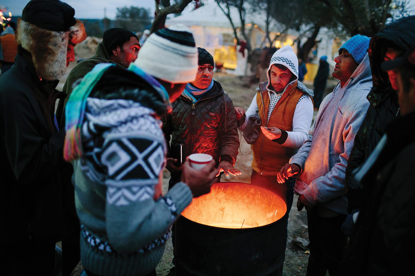 A group of refugees huddle around a fire in a metal trash can.