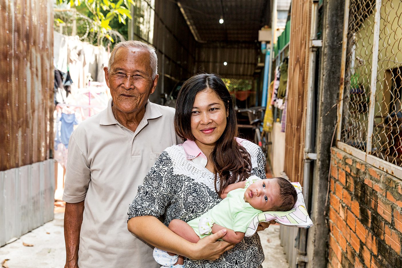 A Cambodian woman holds her baby outside their home. Her father stands behind them.