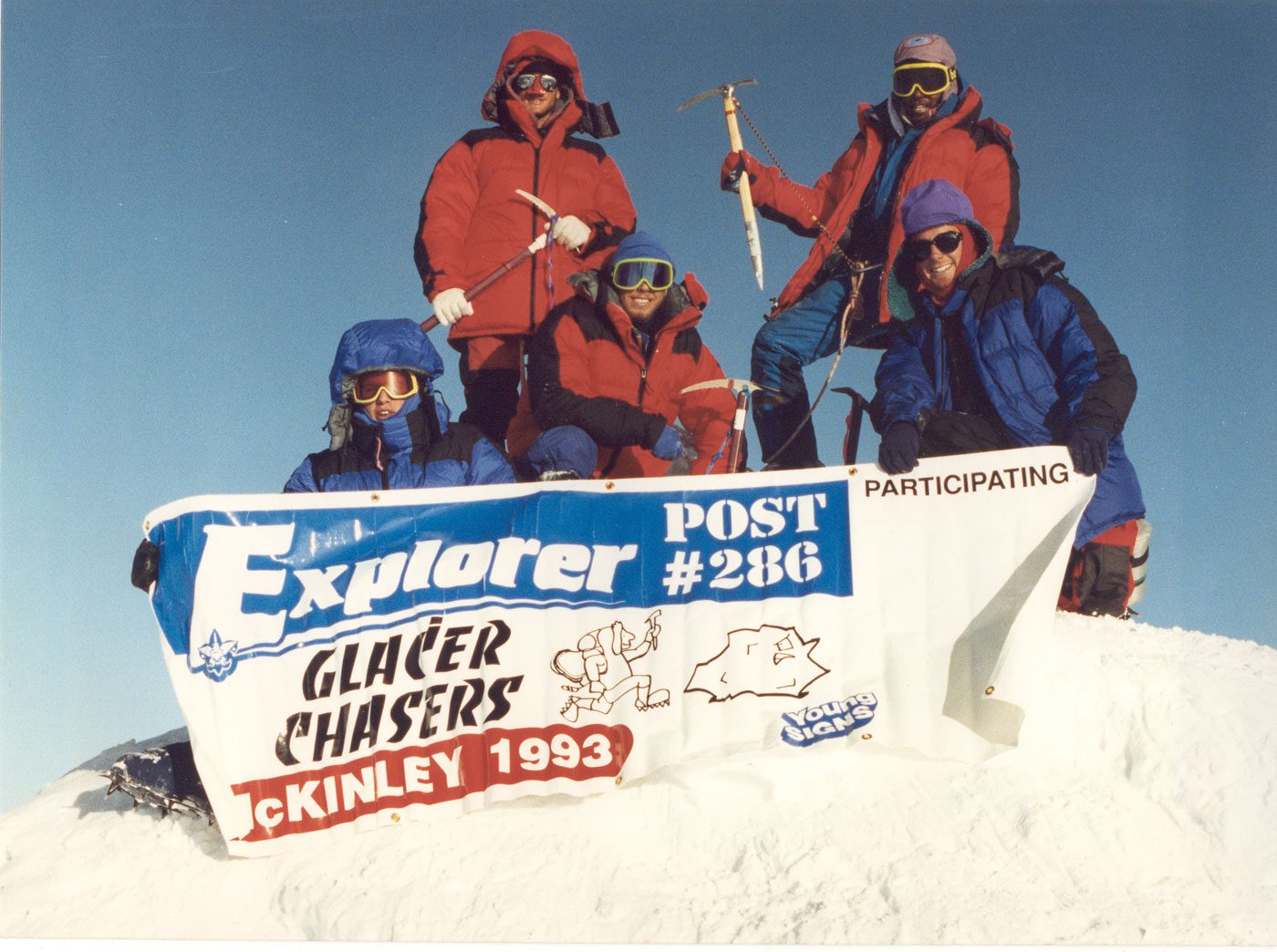 Stacy Taniguchi stands atop Denali with four Boy Scouts. They hold a vinyl sign up with their Scout post number and troop name.