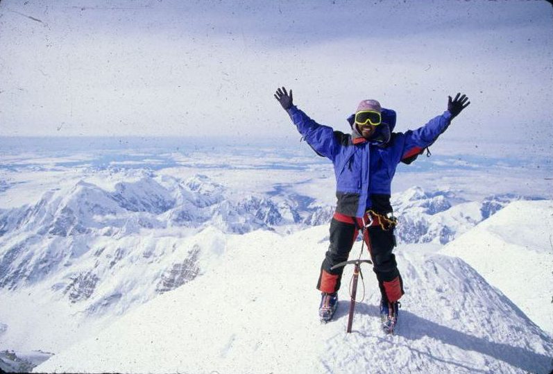 Stacy Taniguchi stands on the summit of Denali with his arms in the air.