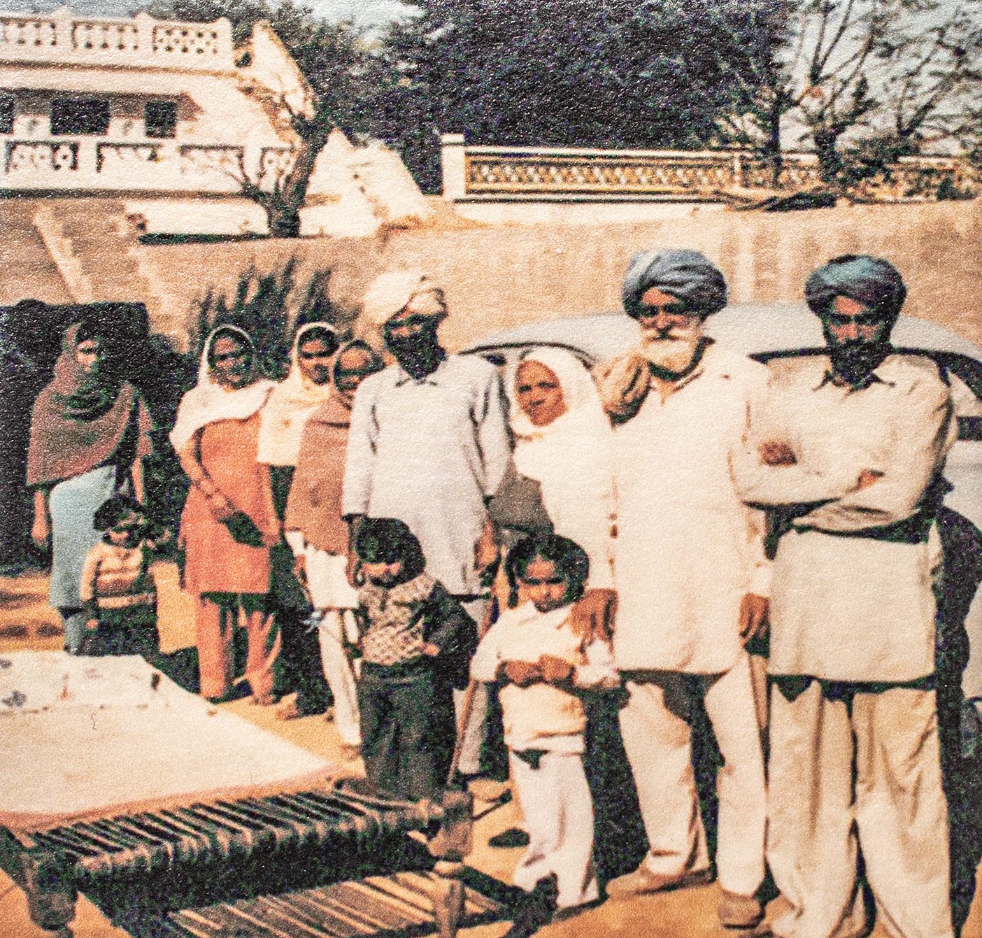 An indian family in traditional Indian garb stands in front of their ancestral home and an old-fashioned car. There is a weaving loom on the ground in front of them.