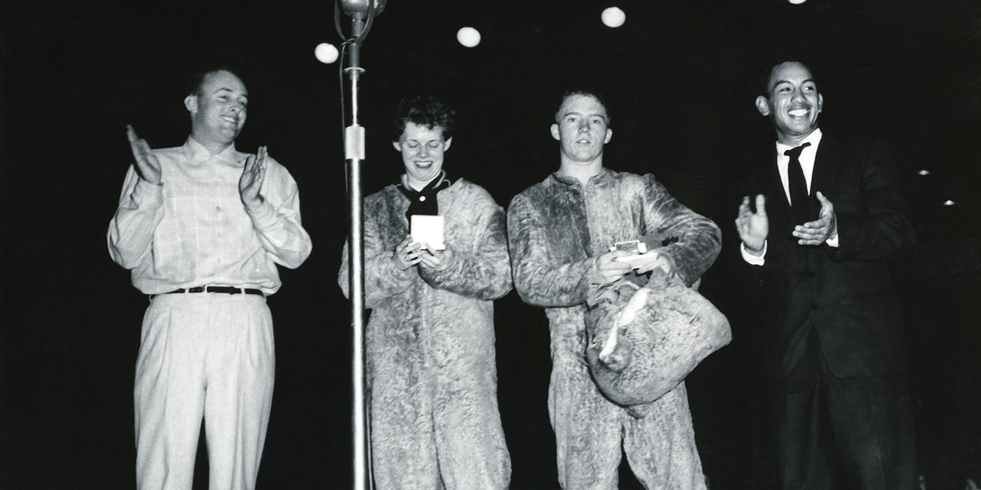 Four people, two of them in Cosmo costume, stand on the playing floor of the Smith Fieldhouse in 1956.