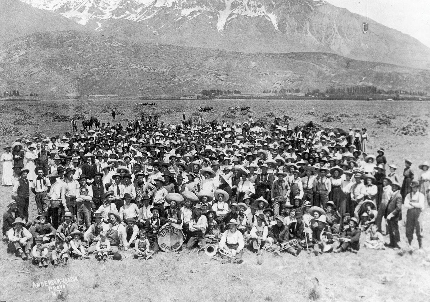 A black and white photo of a large group of students from 1907 dressed as cowboys after clearing out sagebrush from Orem. They pose in a large section of empty land in front of a mountain.