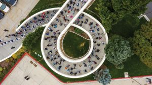 A top down view of the pedestrian ramp near the Marriott Center. It's dotted with people and at the bottom, leading the procession, is the BYU Cougar Marching Band.