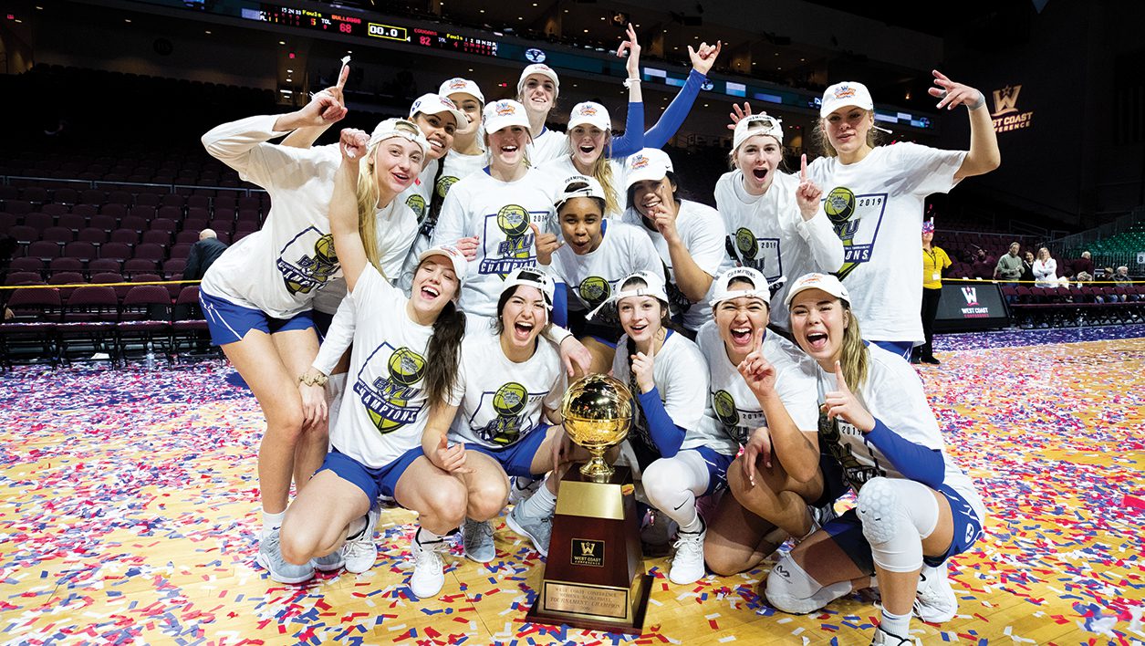 The BYU Women's basketball team gathers in front of the WCC trophy on a confetti-covered court.