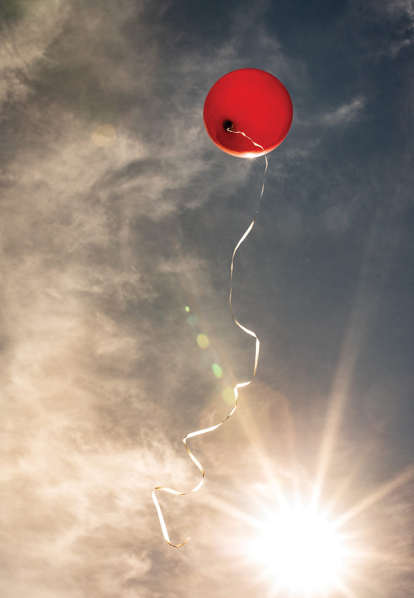 A red balloon with a string attached ascends into a sunlit but cloudy sky.