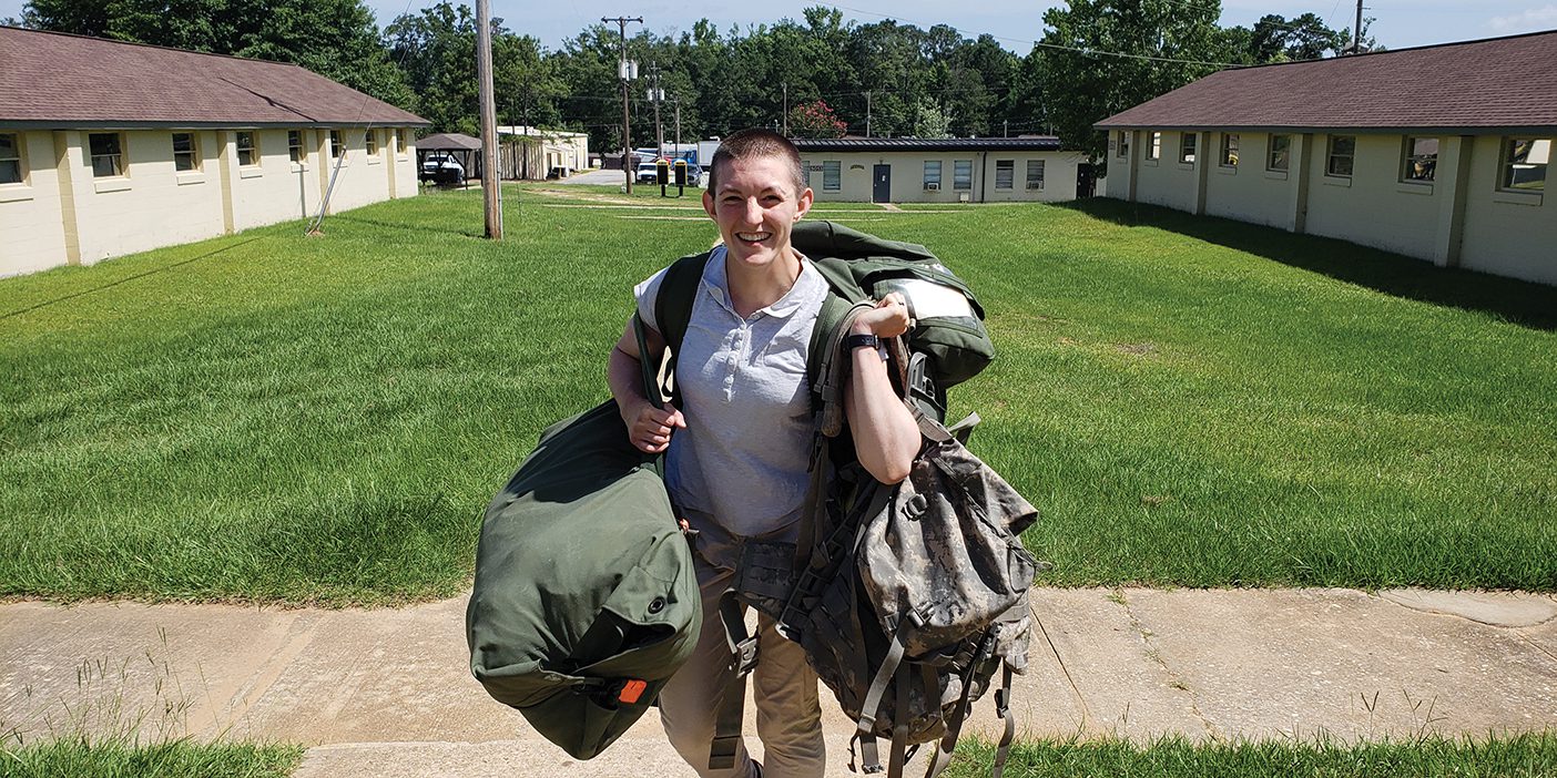 Anna Hodge stands in the middle of a courtyard, heavy canvas bags in both her hands and dangling off her shoulders. She is smiling broadly at the camera.