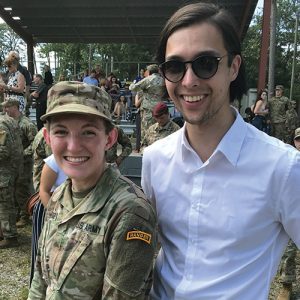 Anna Hodge and her husband Matthew stand close together outside. Anna is in military uniform, as are the people in the background.