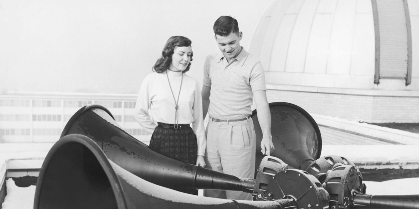 Old photo of a man and a woman looking at large horns to give off the sound of a bell on a rooftop.