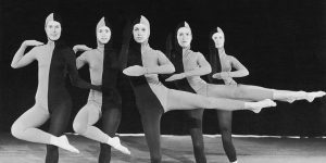 Five dancers dressed in full body tight suits split in half with gray and black take dancing poses for the camera.