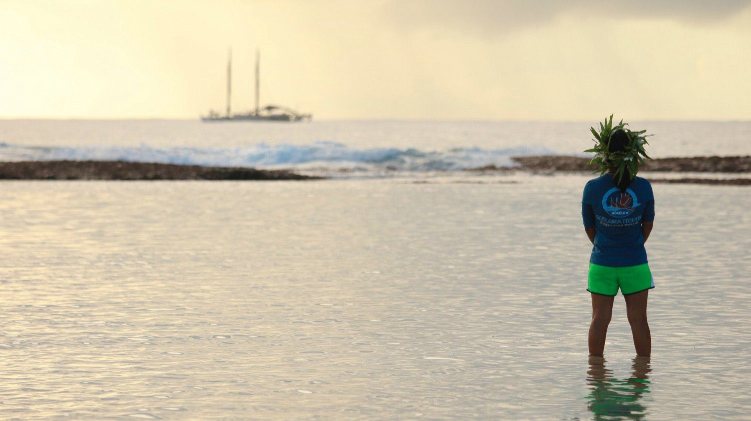 Furuto stands in the ocean looking at a sailboat in the water