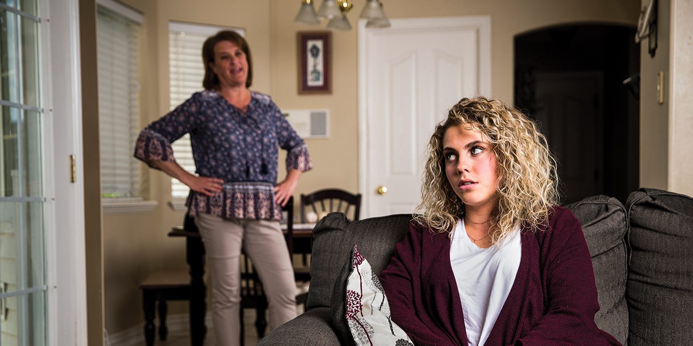 teenage girl sitting on a sofa rolls her eyes at her mother, hands on hips, talking to her in the background.