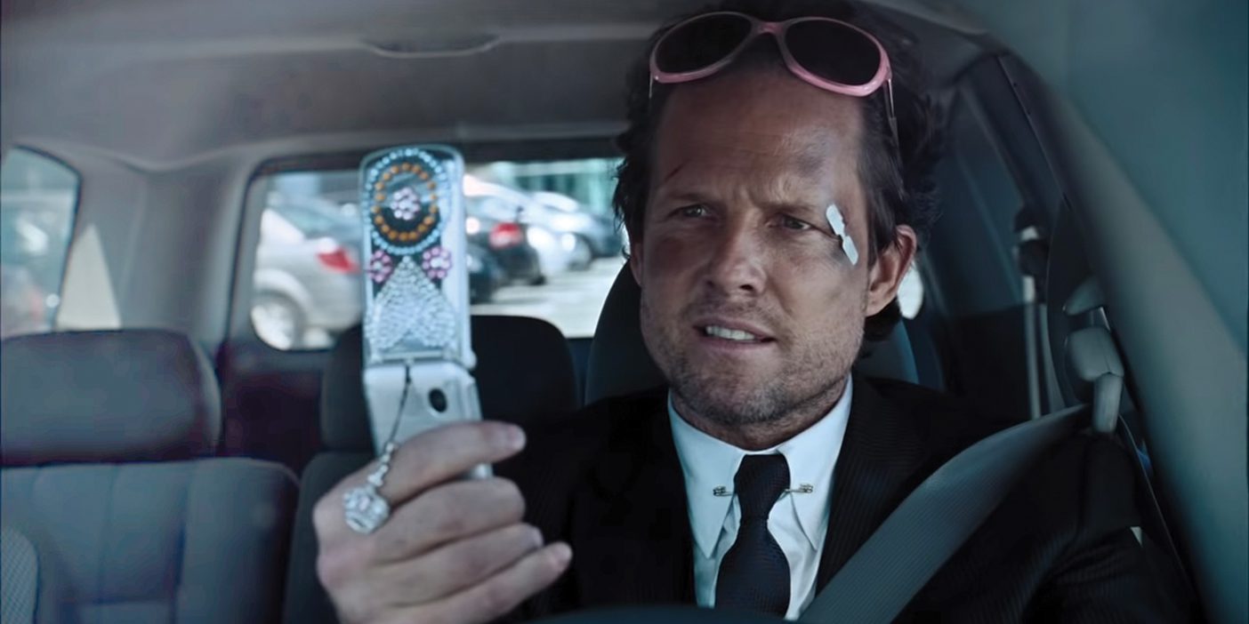 Mayhem, a character from AllState Insurance commercials, wears a black suit and tie and pink sunglasses on his foreheard. He holds a broken phone and has a bruised, scratched face.