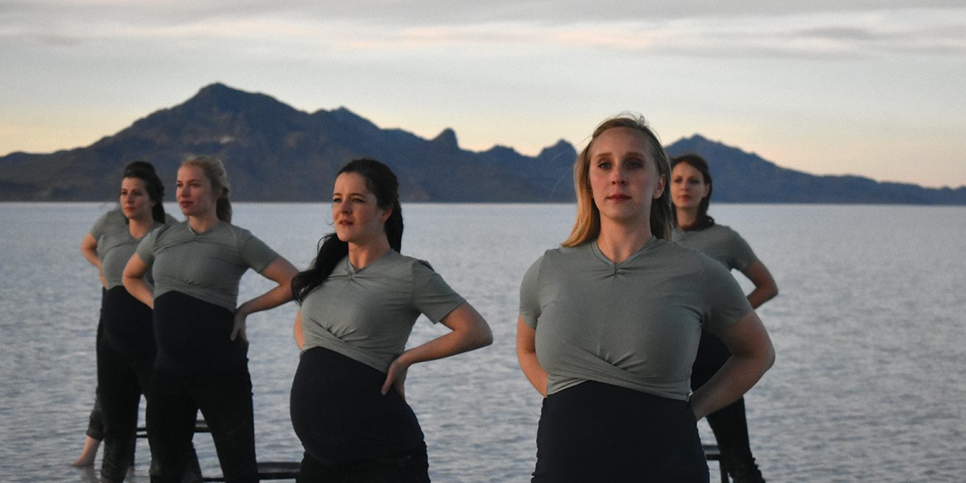 Five pregnant dancers stand looking outward with their hands on their hips.