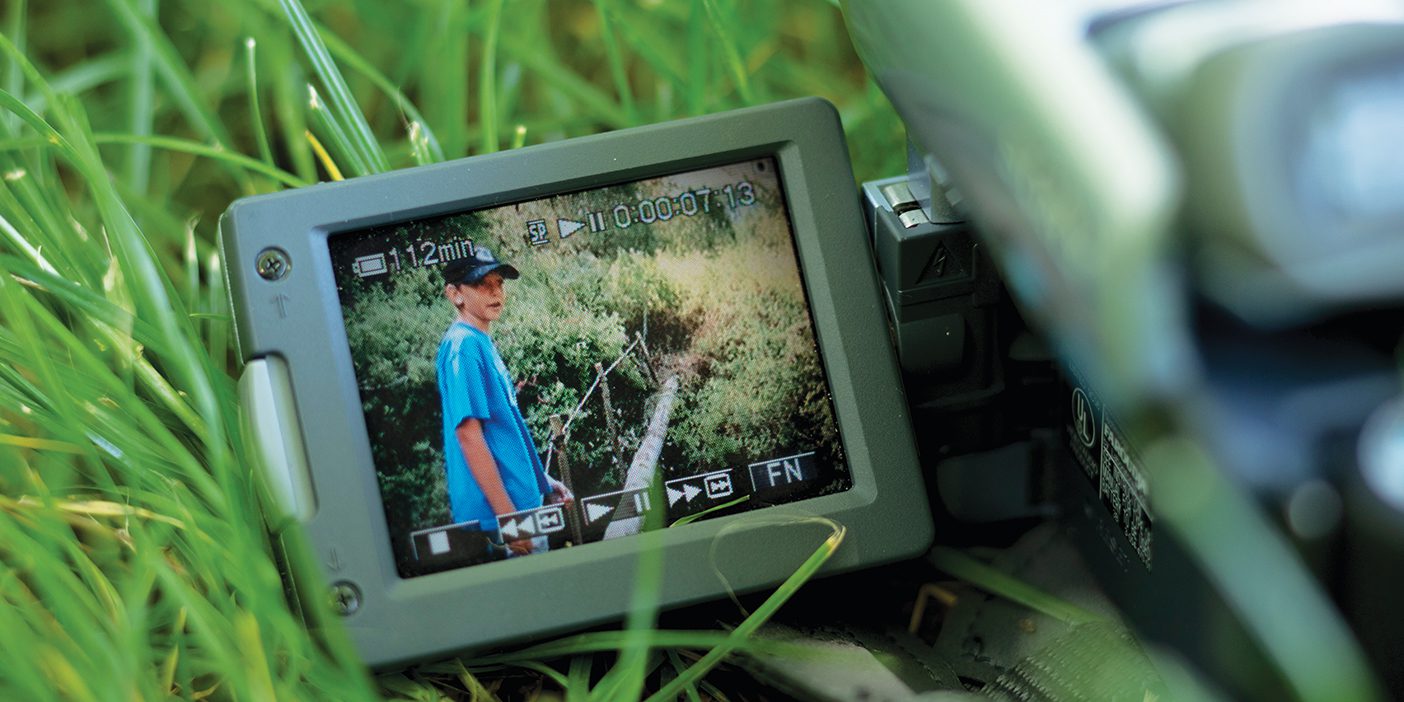 A video camera lies on the grass. On the screen of the camera, a young boy stands next to a wooden bridge stretching across a small stream.
