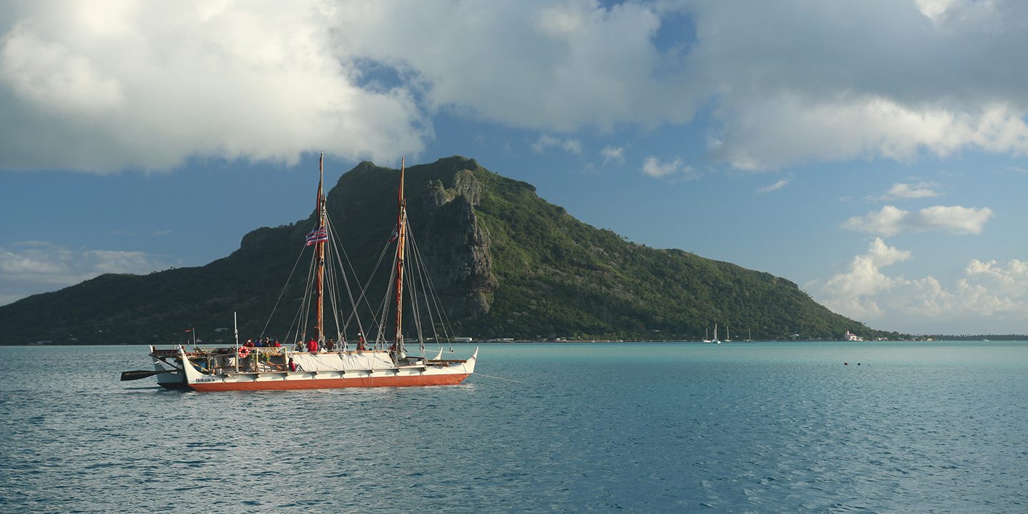 The canoe sails past a small mountainous island in the ocean.