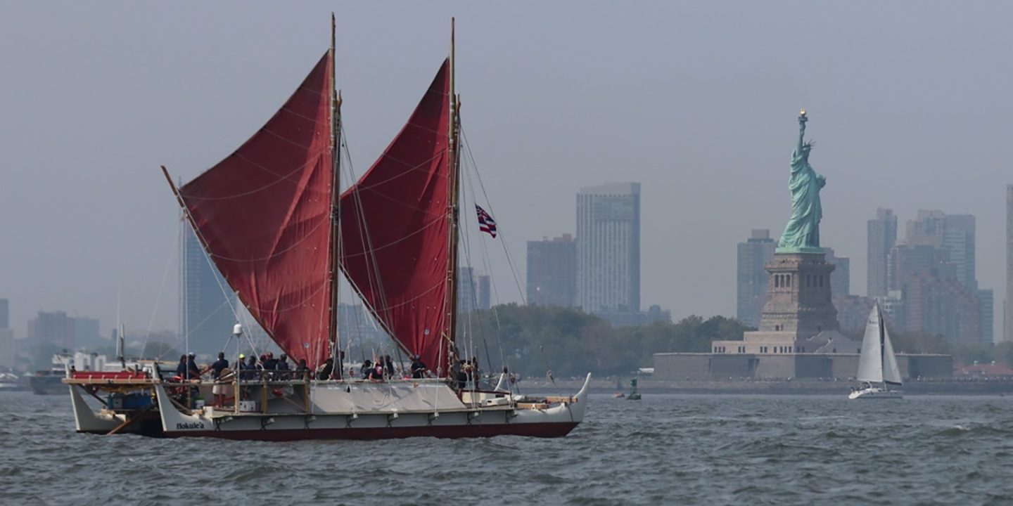 The voyaging canoe sails with the New York City skyline in the background featuring the Statue of Liberty