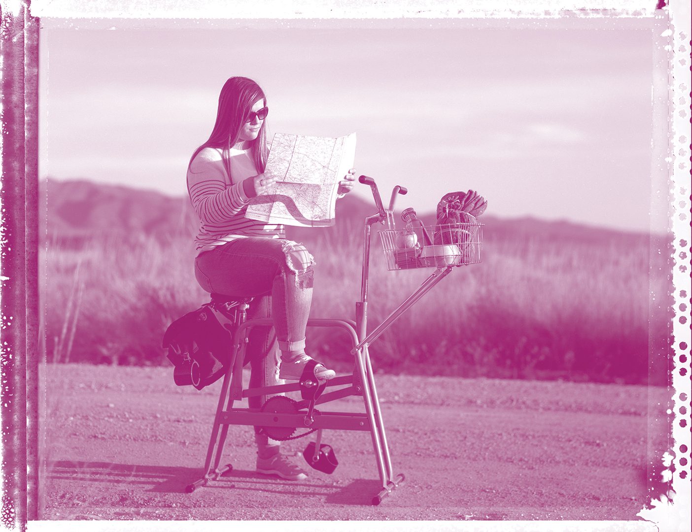 Girl looking at a map and riding a stationary bike on a country roadside. Depicts futility of getting somewhere.