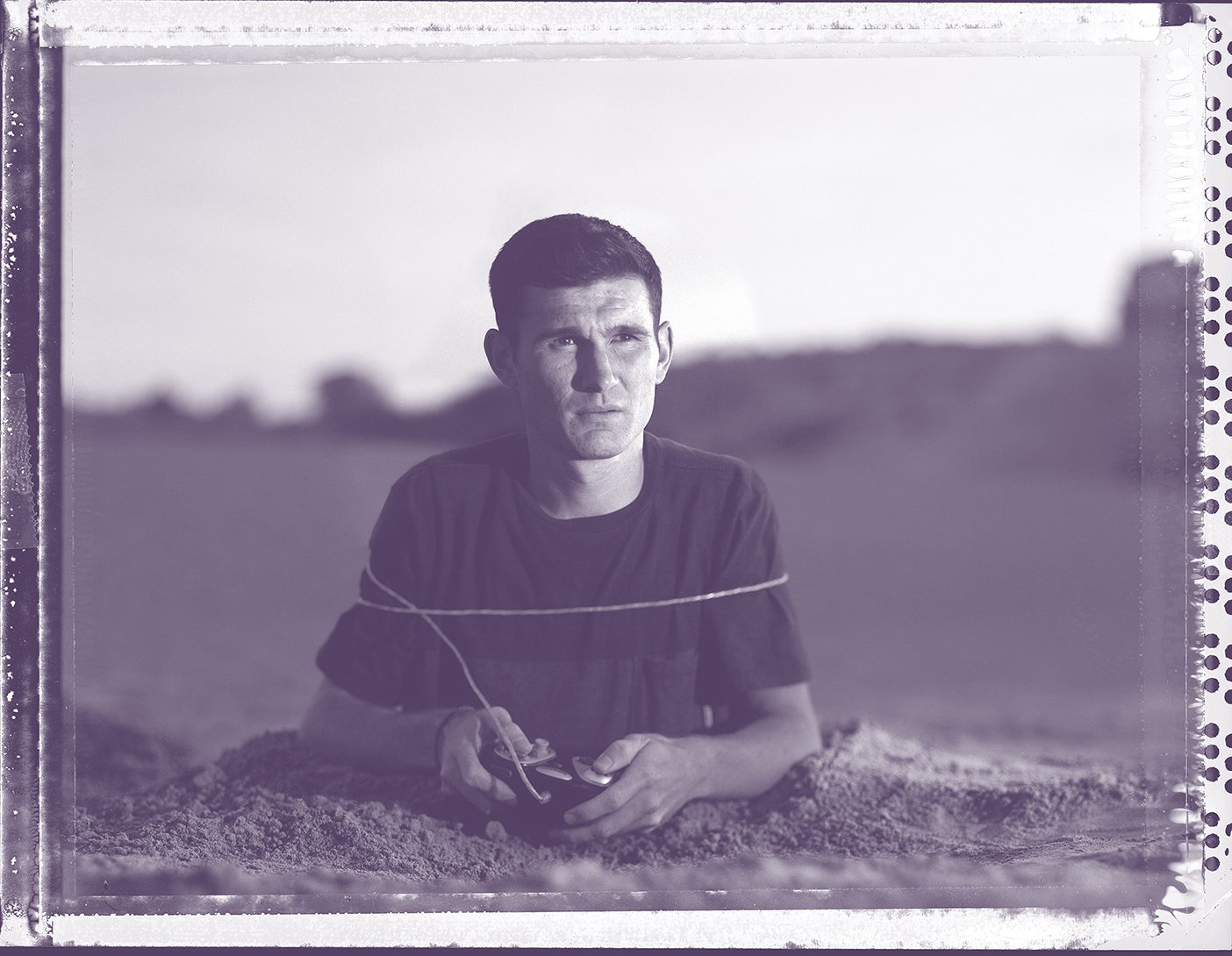 Young adult playing a video game while buried up to his waist in sand.