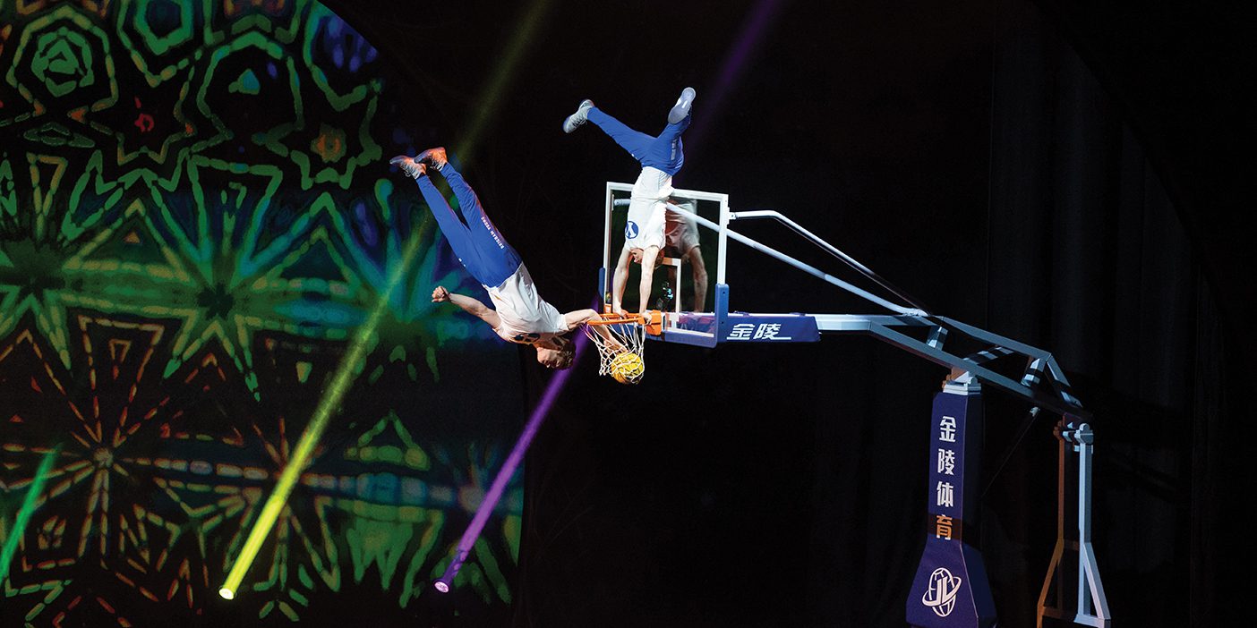 Two dunk team members flip acrobatically above a basketball hoop.