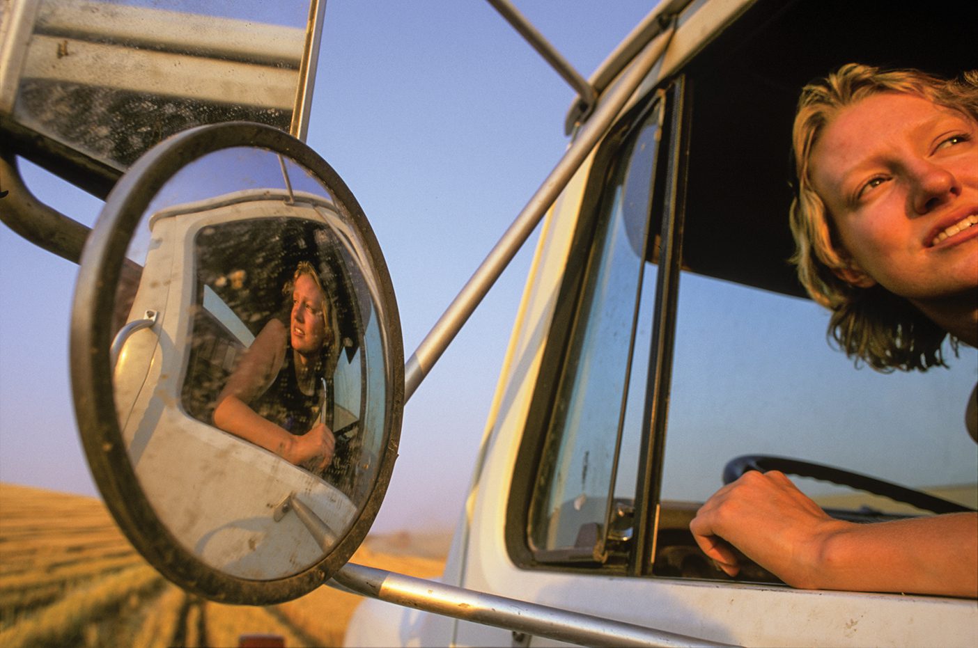 A person leans out a car window and their reflection can be seen in the large side mirror.