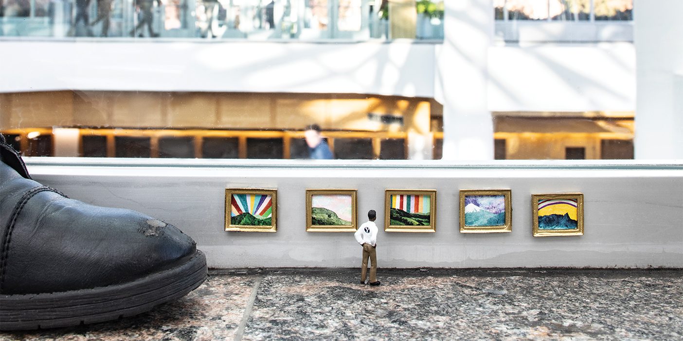 A human-sized foot stands next to the art display, showing just how tiny it is; the miniature admirer is looking at a gallery of thumbnail-sized art.