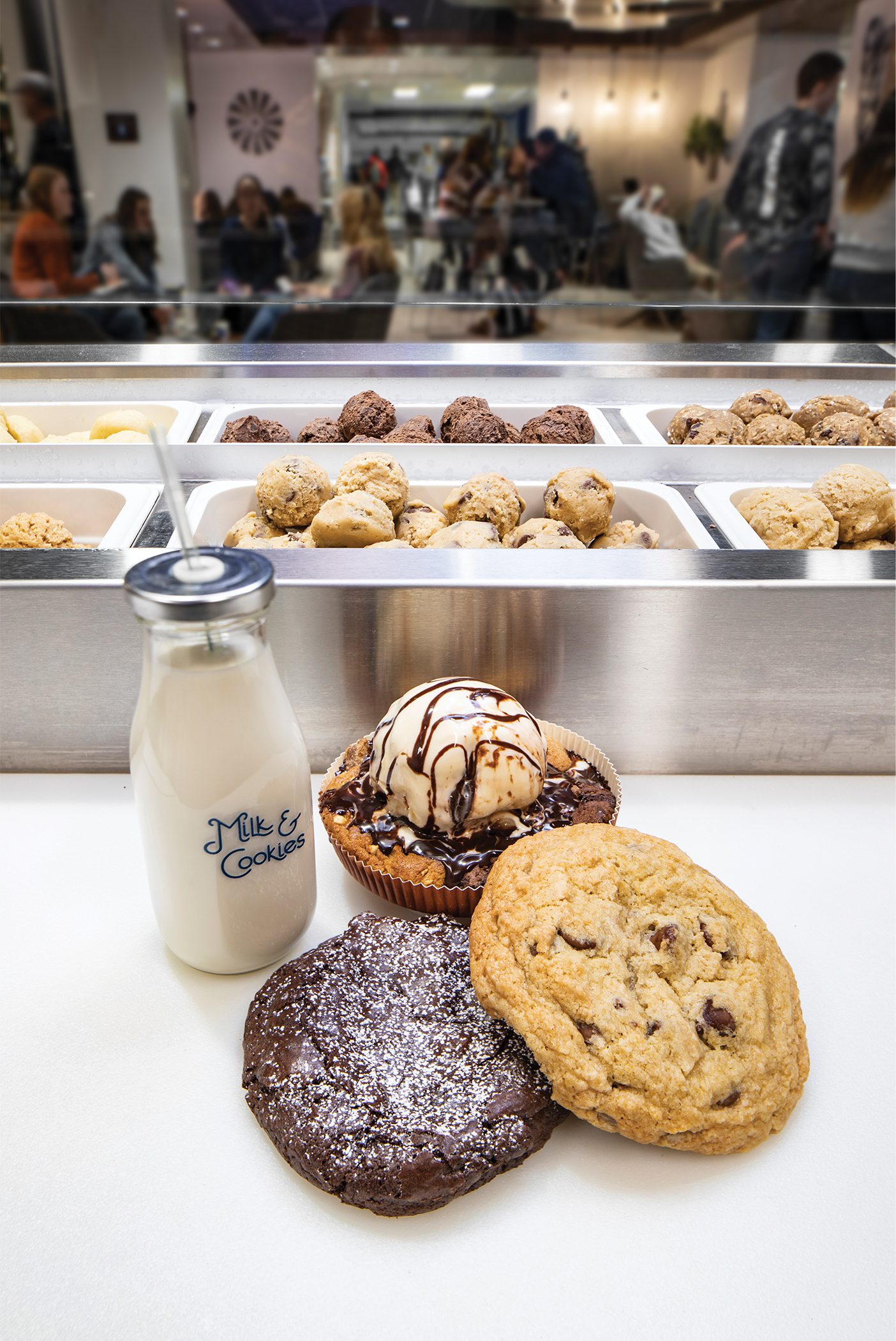 A jar of milk sits on a counter alongside two large cookies and a Cosmookie.