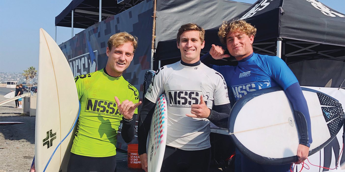 Three smiling Brigham Young Surf Team members hold surf boards and make the hang loose hand gesture.