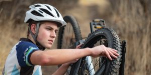 A young man wearing a bicycle helmet and other gear repairs the flat tire on his mountain bike.